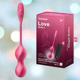 The image features the Satisfyer Love Birds 2 device in pink, placed next to its packaging. The pink box emphasizes key features like Bluetooth connectivity, vibration capabilities, and a full silicone design, making it ideal for individuals looking for impactful pelvic floor training with vibrating Ben Wa balls functionality.